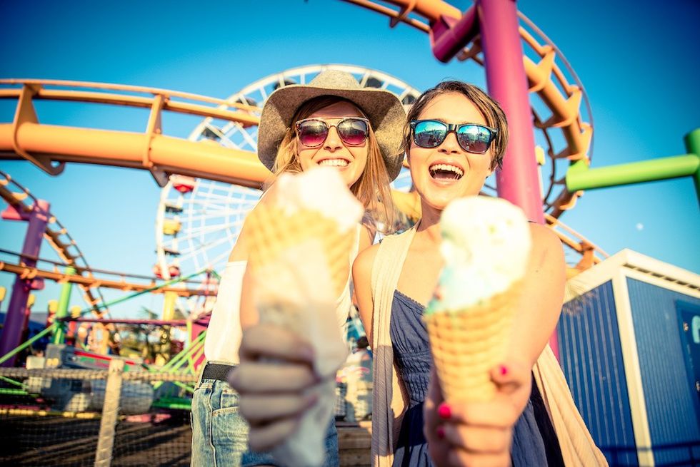 Two women enjoy ice cream at an amusement park with a roller coaster and Ferris wheel.