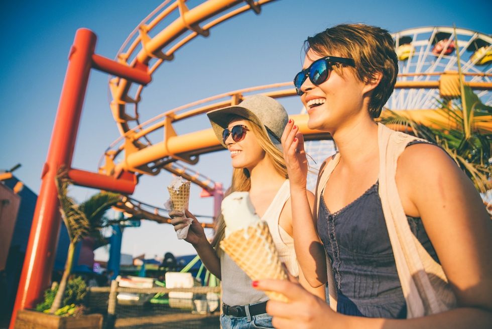 Two women enjoying ice cream at an amusement park, with a roller coaster in the background.