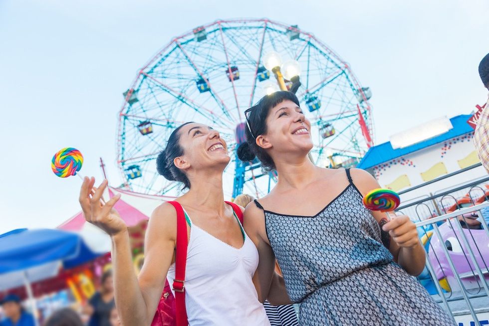 Two women enjoying lollipops at a carnival with a Ferris wheel in the background.