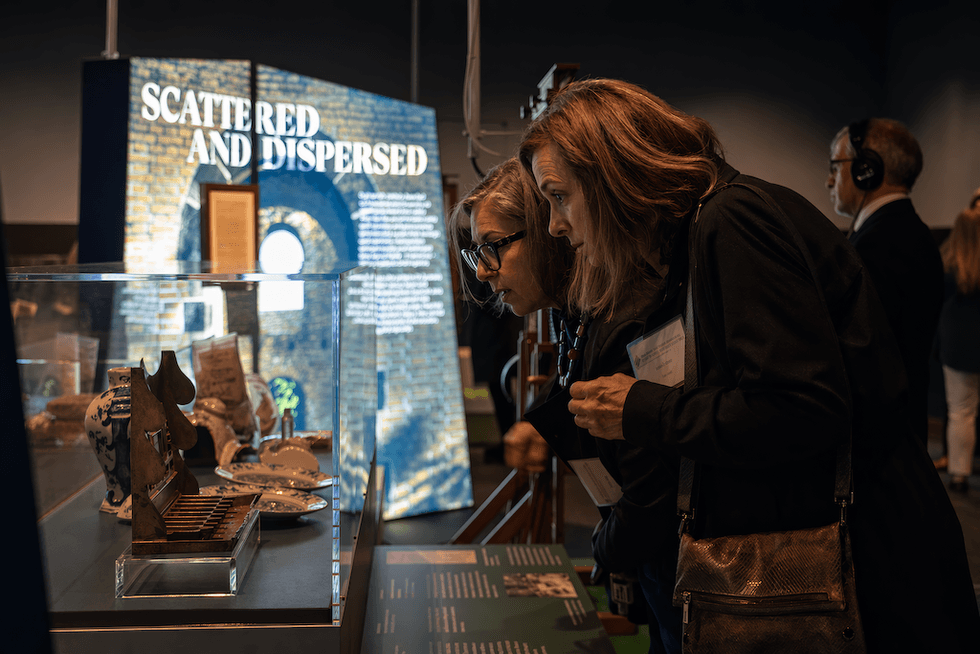 Two women examining museum artifacts behind glass display titled "Scattered and Dispersed."
