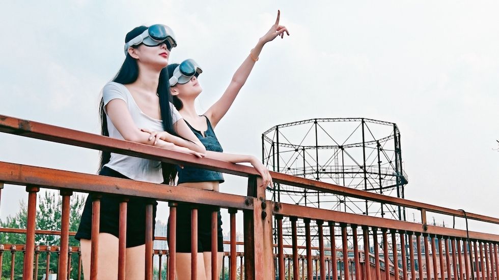 Two women in goggles stand on a bridge, one pointing to the sky.