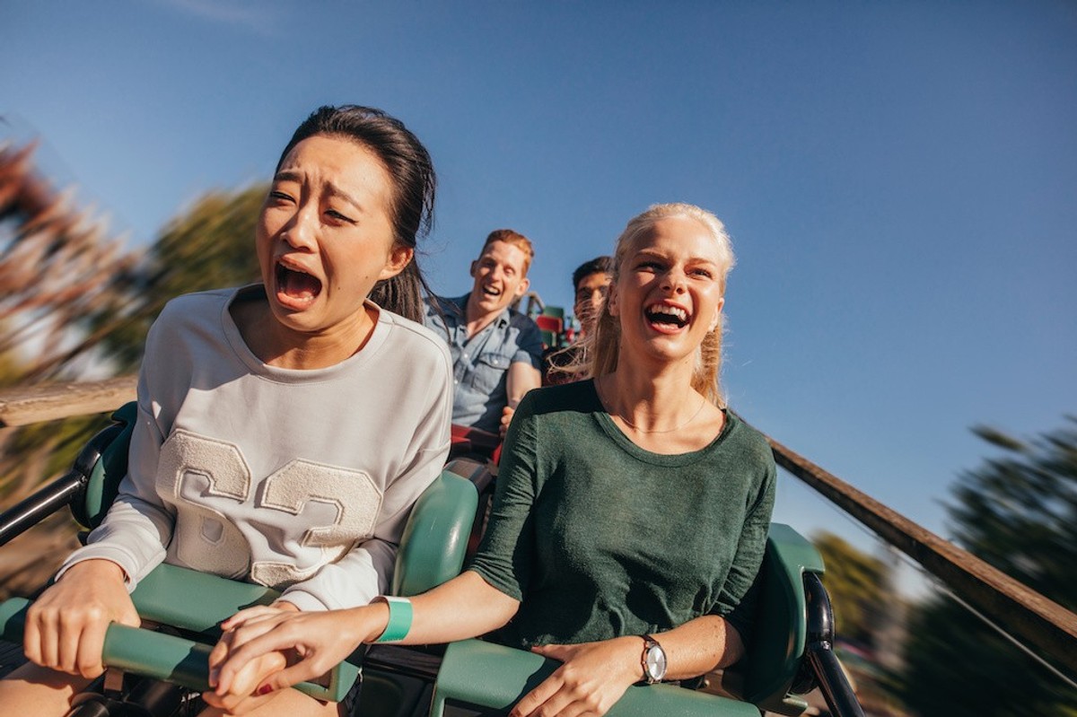 Two women on a roller coaster, one screaming, one laughing, holding hands.