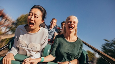 Two women on a roller coaster, one screaming, one laughing, holding hands.
