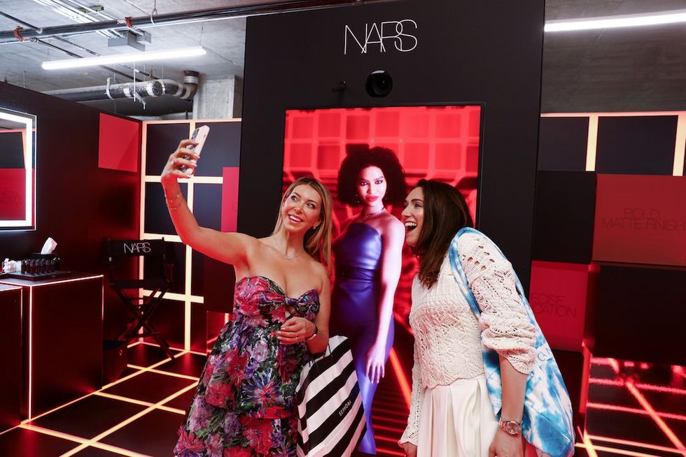 Two women take a selfie at a NARS cosmetics display with vibrant lighting.