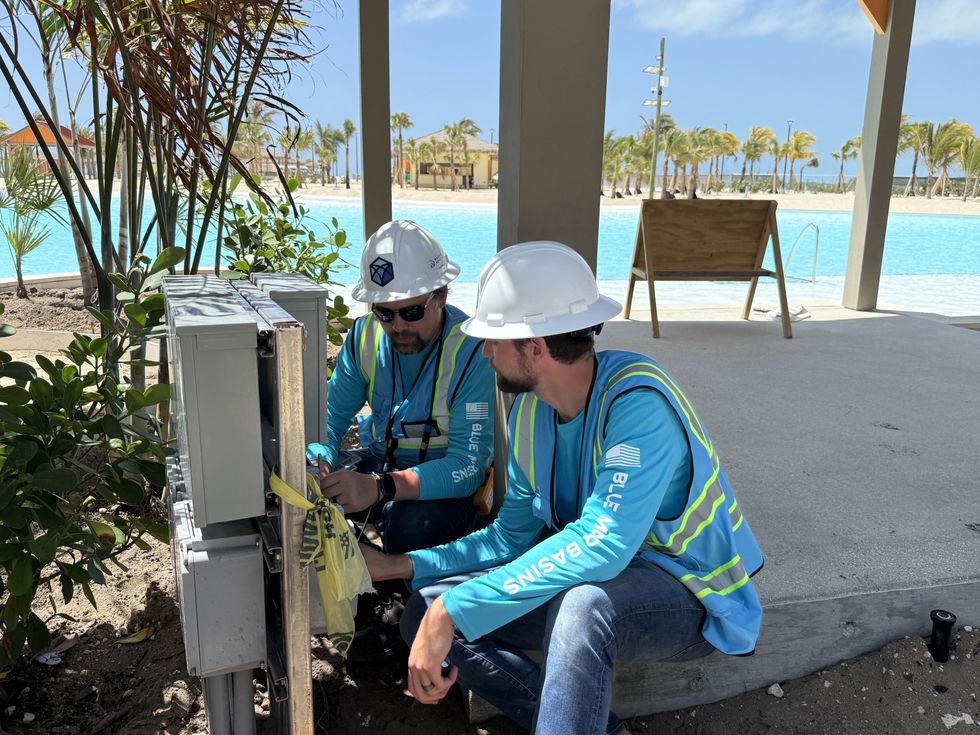 Two workers in hard hats examine an electrical box by a poolside.