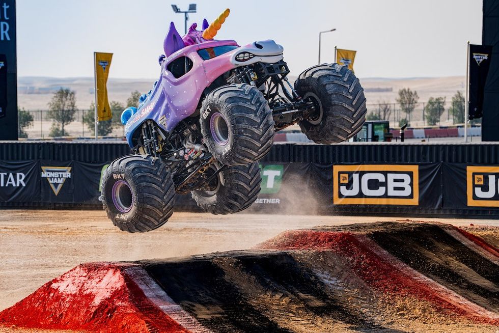 Unicorn-themed monster truck jumps over dirt ramp at an outdoor event.