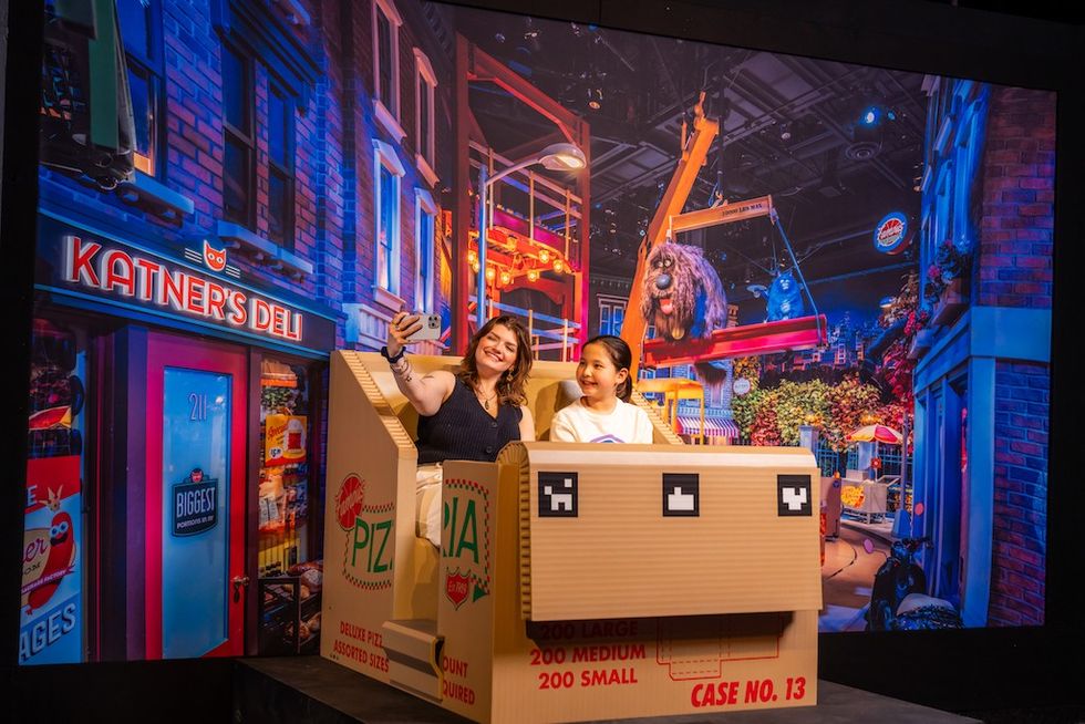 Universal Theme Parks: The Exhibition - two people taking a selfie in a whimsical, colorful set with pizza box props and street backdrop.