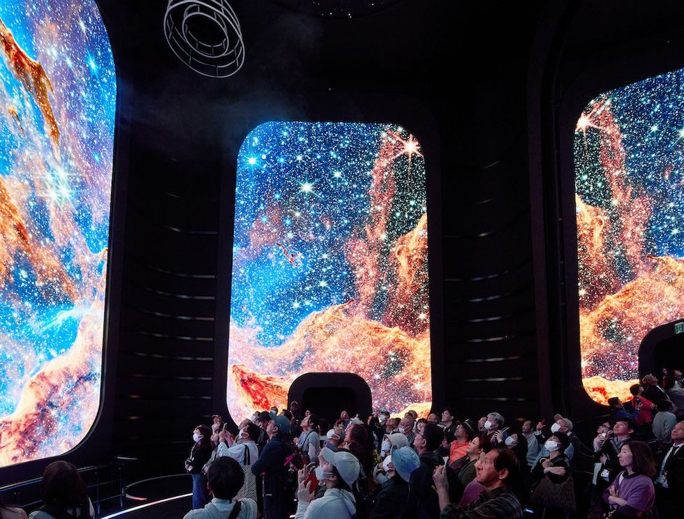 USA Pavilion rocket launch. People gaze at vibrant galaxy visuals on large indoor screens.