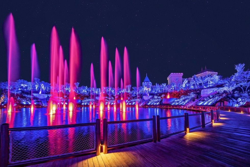 Vibrant purple fountains under a starry night sky, with illuminated trees and a boardwalk.