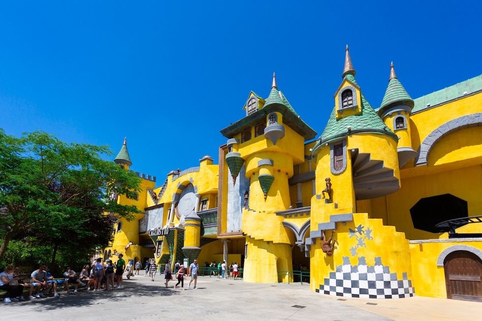 Vibrant yellow building with green turrets under a clear blue sky, people walking nearby.