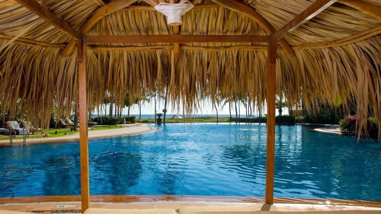 View from under a thatched cabana by a pool, with a distant ocean horizon.