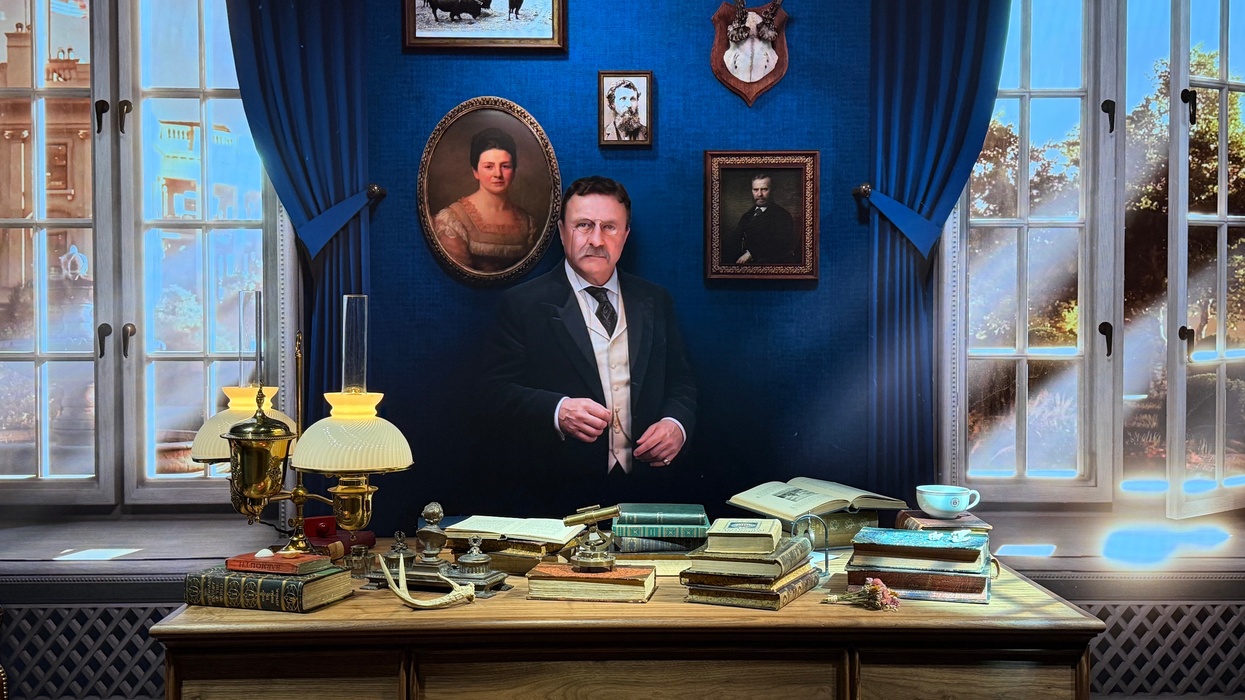 Vintage study with Theodore Roosevelt, antique desk, books, lamps, and portraits on a blue wall.