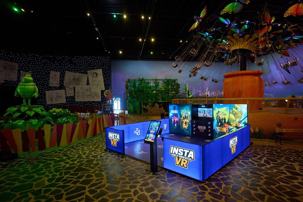 VR gaming booth in a colorful amusement park with a swing ride and green character nearby.