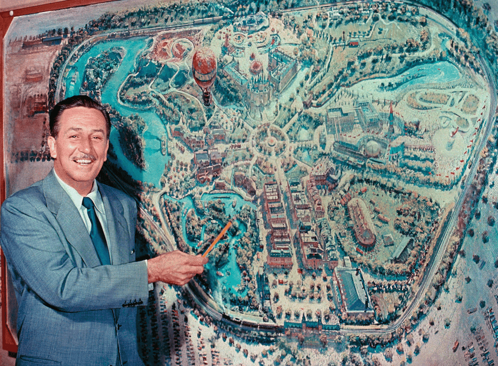 Walt Disney pointing at an illustrated map of a theme park with a pointer.