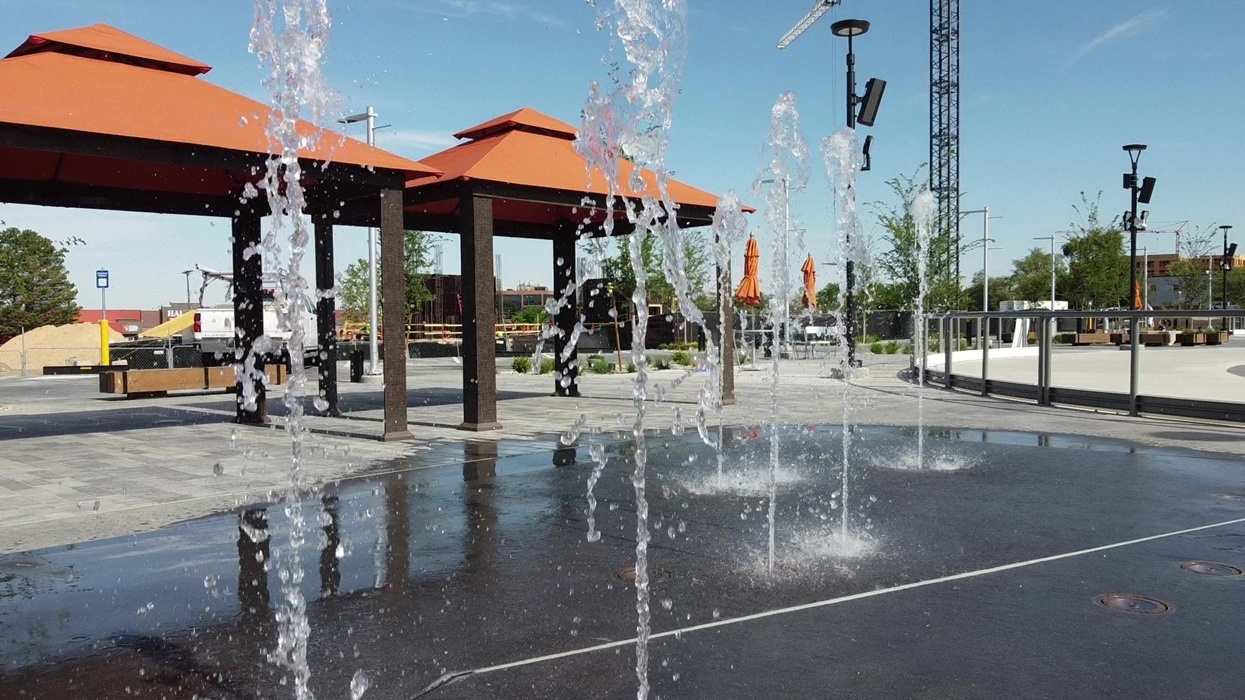 Water fountains in a plaza with orange-roofed shelters and a clear blue sky.
