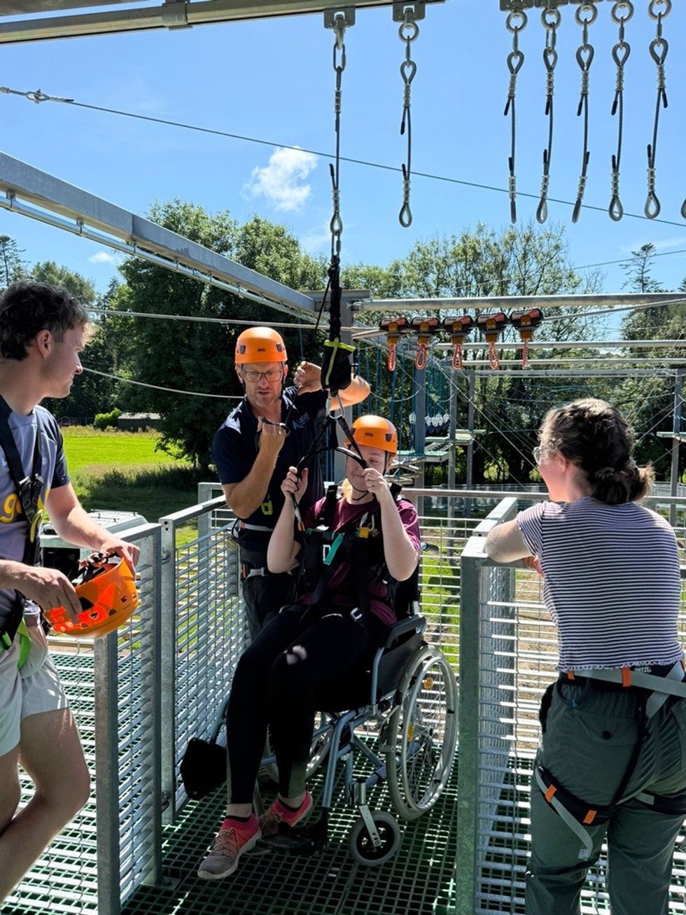 Wheelchair user preparing for a zip line with assistance on a sunny day.