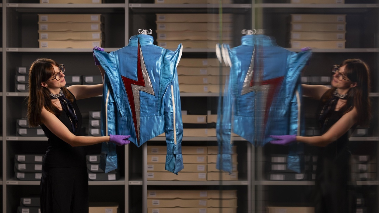 Woman holding a blue jacket belonging to David Bowie with red lightning bolt, reflected in a glass case