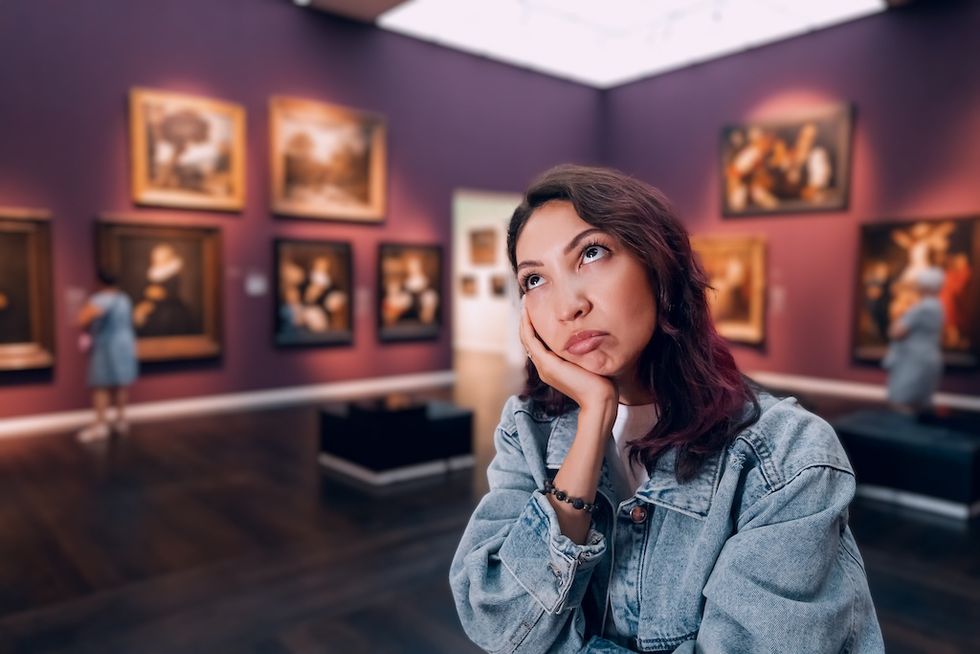 Woman in art gallery looking bored, resting head on hand, surrounded by paintings.