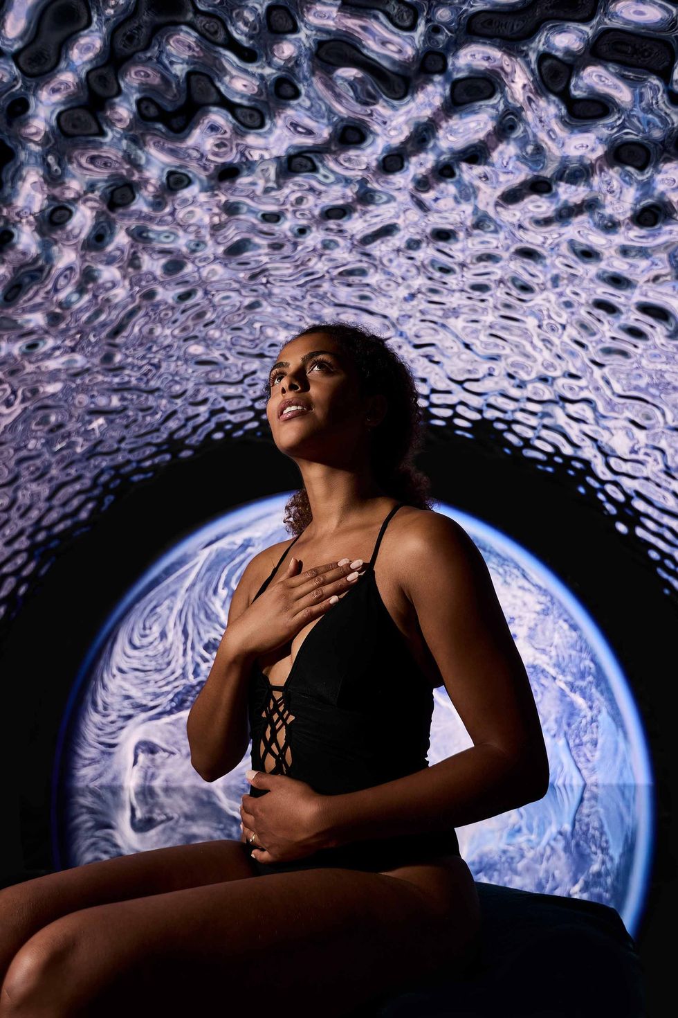 Woman in black dress gazes upward, cosmic swirl pattern overhead, Earth-like backdrop.