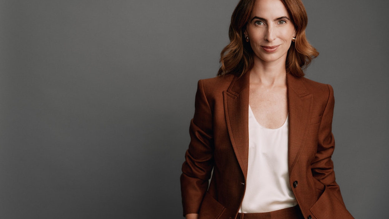 Woman in brown suit and white blouse, smiling against a gray background.