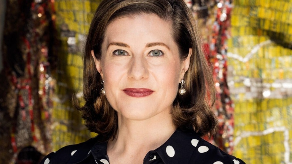 Woman in polka dot blouse stands against a colorful mosaic backdrop.