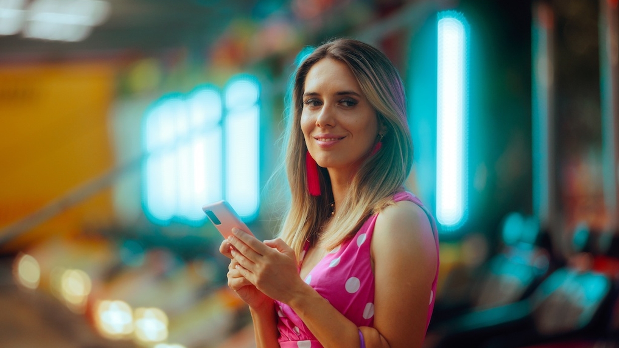 Woman in polka dot dress smiles, holding phone, with neon lights in the background.