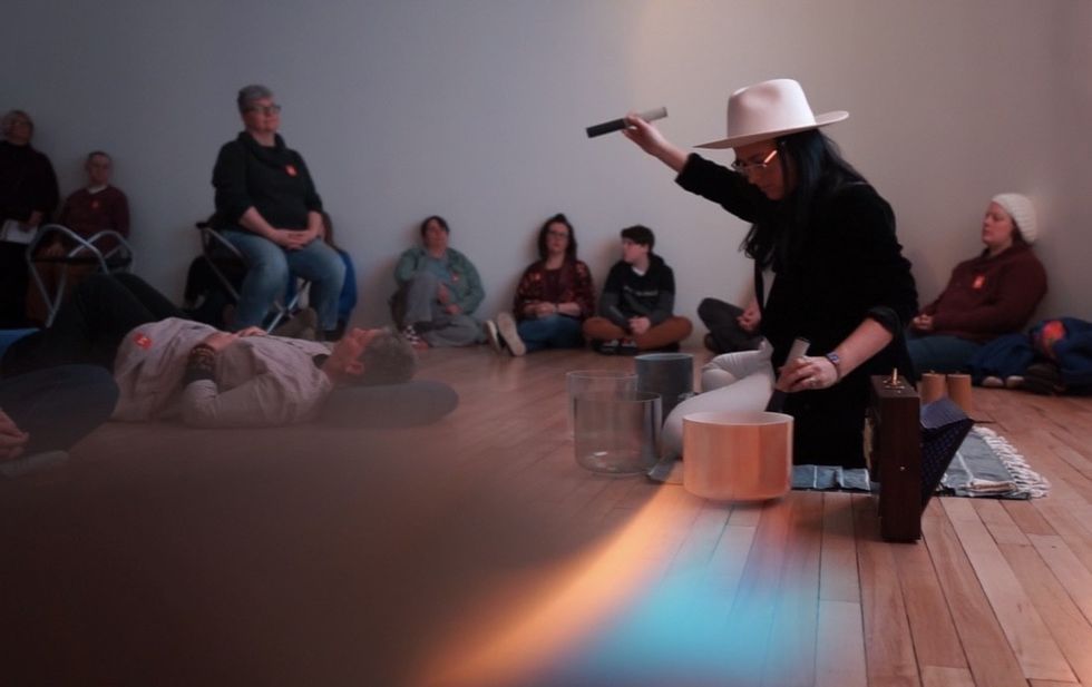 Woman in white hat conducting sound healing session, surrounded by seated people.