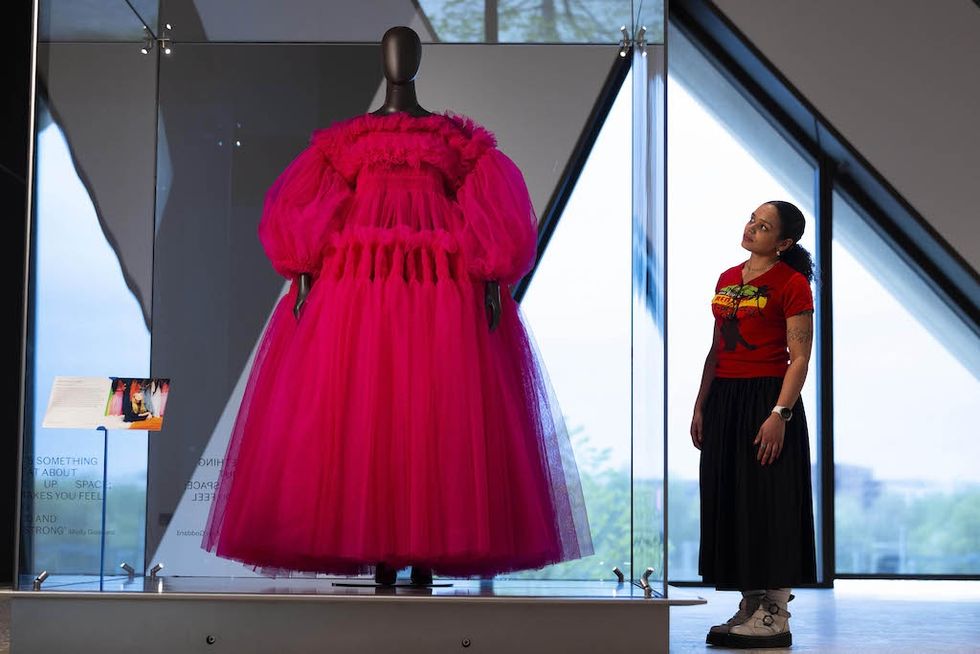 Woman observing vibrant pink dress in glass display case at V&A East