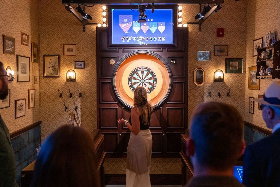 Woman playing darts in a cozy, vintage-style room with an audience watching.