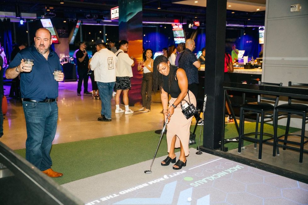Woman playing mini-golf indoors with a drink, while others socialize in the background.