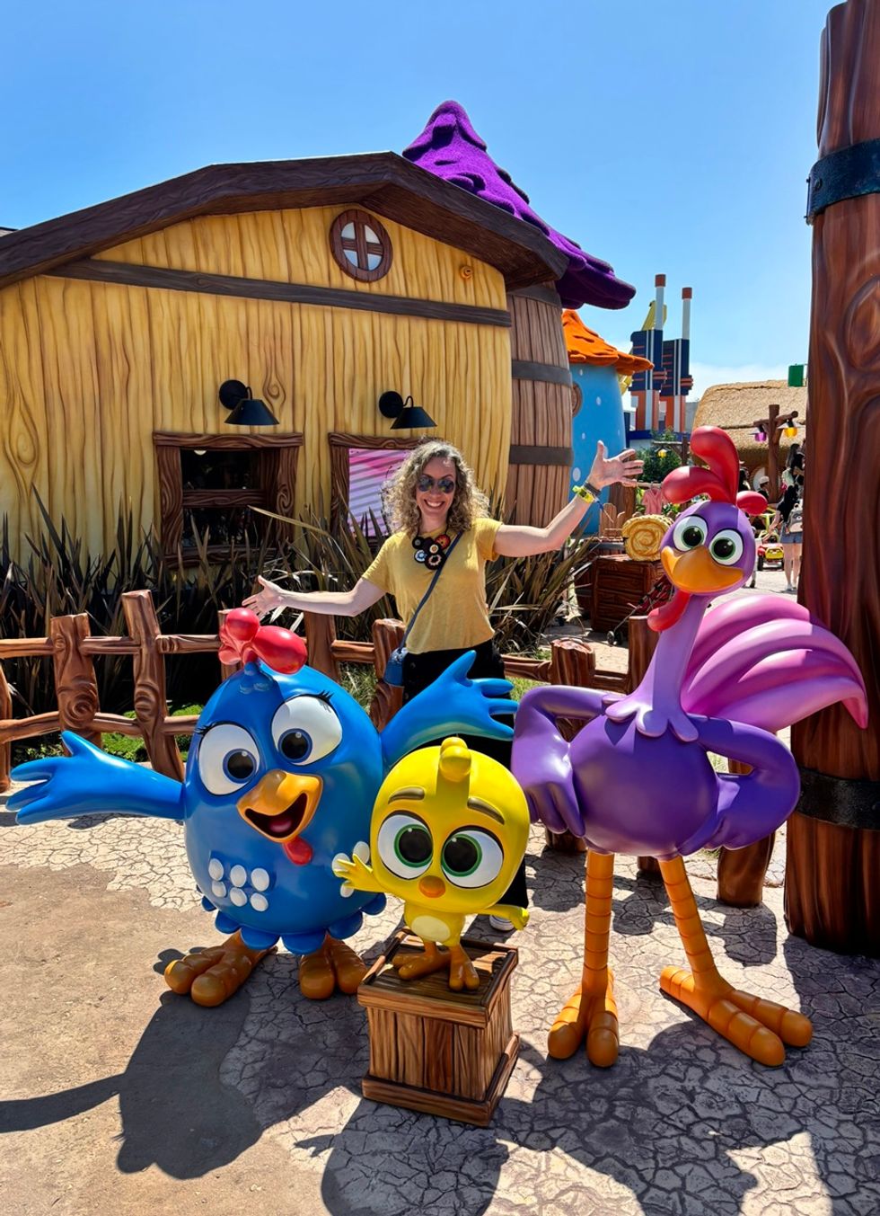 Woman posing with colourful cartoon bird statues in a whimsical outdoor setting at Vila da Galinha Pintadinha at Beto Carrero World