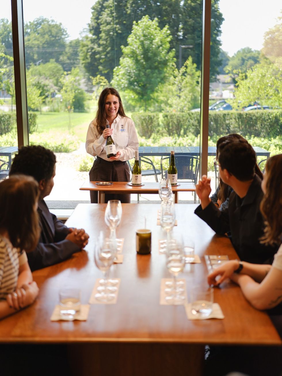Woman presenting wine to a seated group in a bright room with large windows.