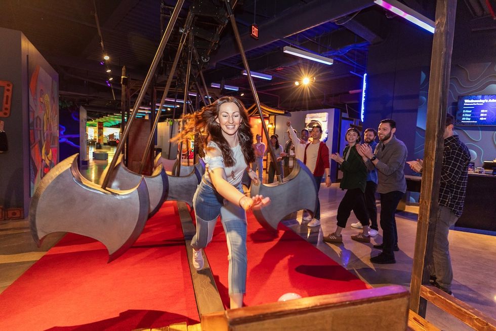 Woman running through swinging axes in a funhouse, with people cheering her on.
