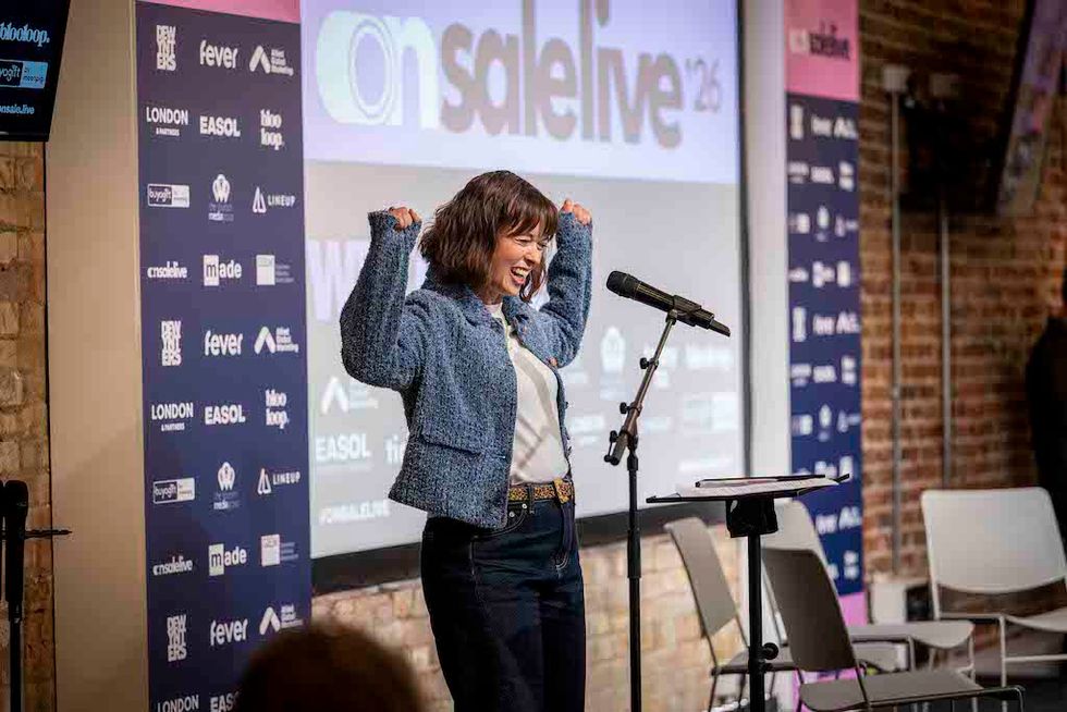 Woman smiling and raising arms while speaking at a podium in front of a conference banner.