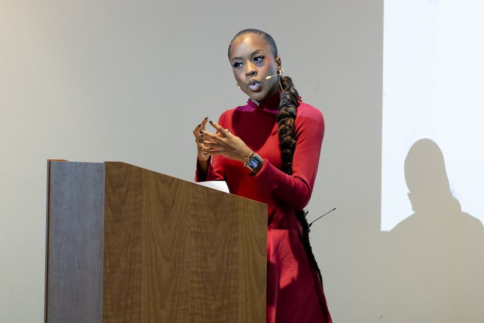 Woman speaking at a podium, wearing a red dress and a long braided ponytail.