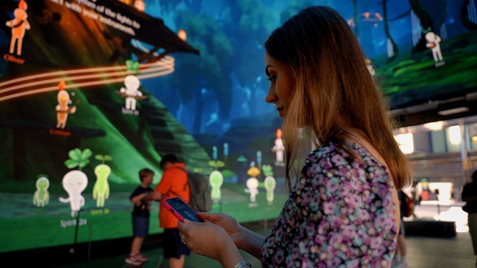 Woman using phone in front of animated forest display in a dimly lit room.