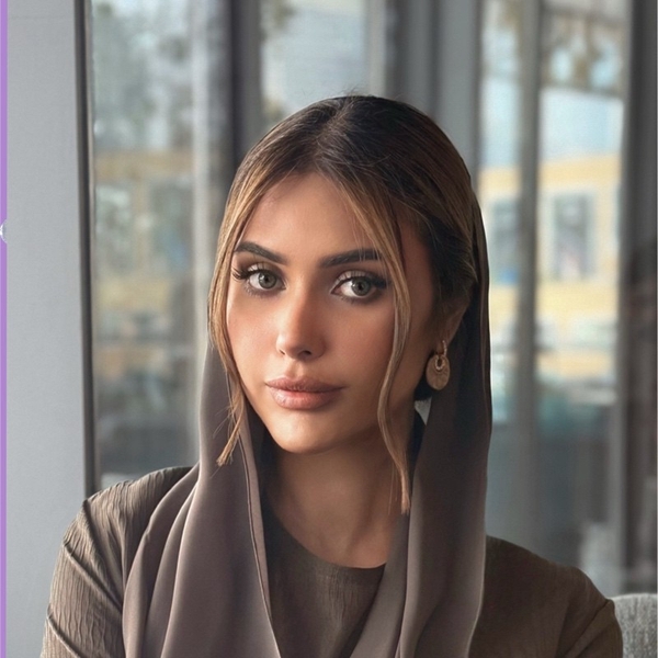 Woman with long hair and earrings wears a headscarf, sitting indoors by a window.