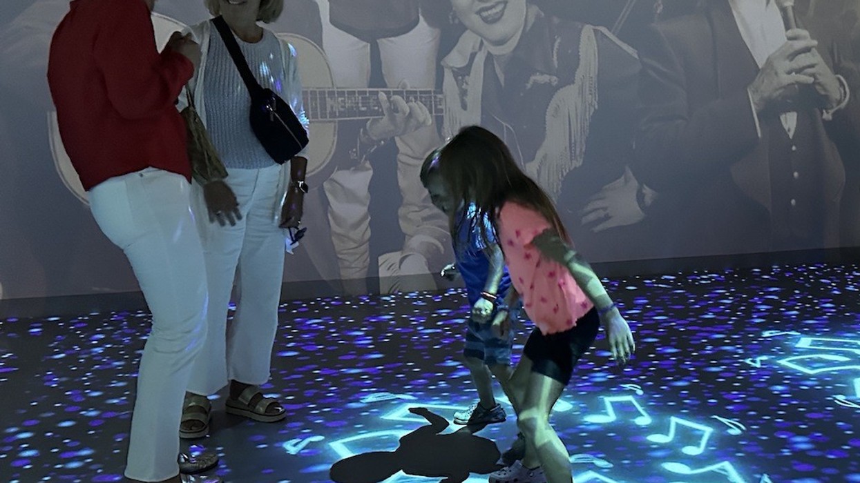 Women and children dance on a lighted floor with music notes; musicians on the wall background.