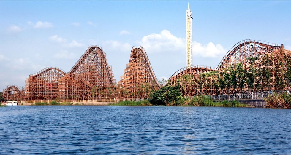 Wooden roller coaster by a lake under a blue sky with a tall tower in the background.