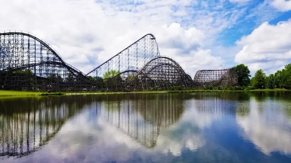 Wooden roller coaster reflected in a calm lake under a partly cloudy sky.