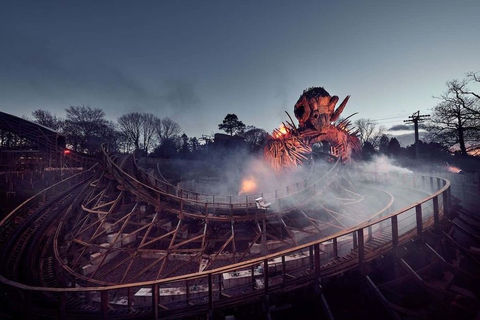 Wooden roller coaster, Wickerman, with a fiery sculpture at dusk, surrounded by mist.