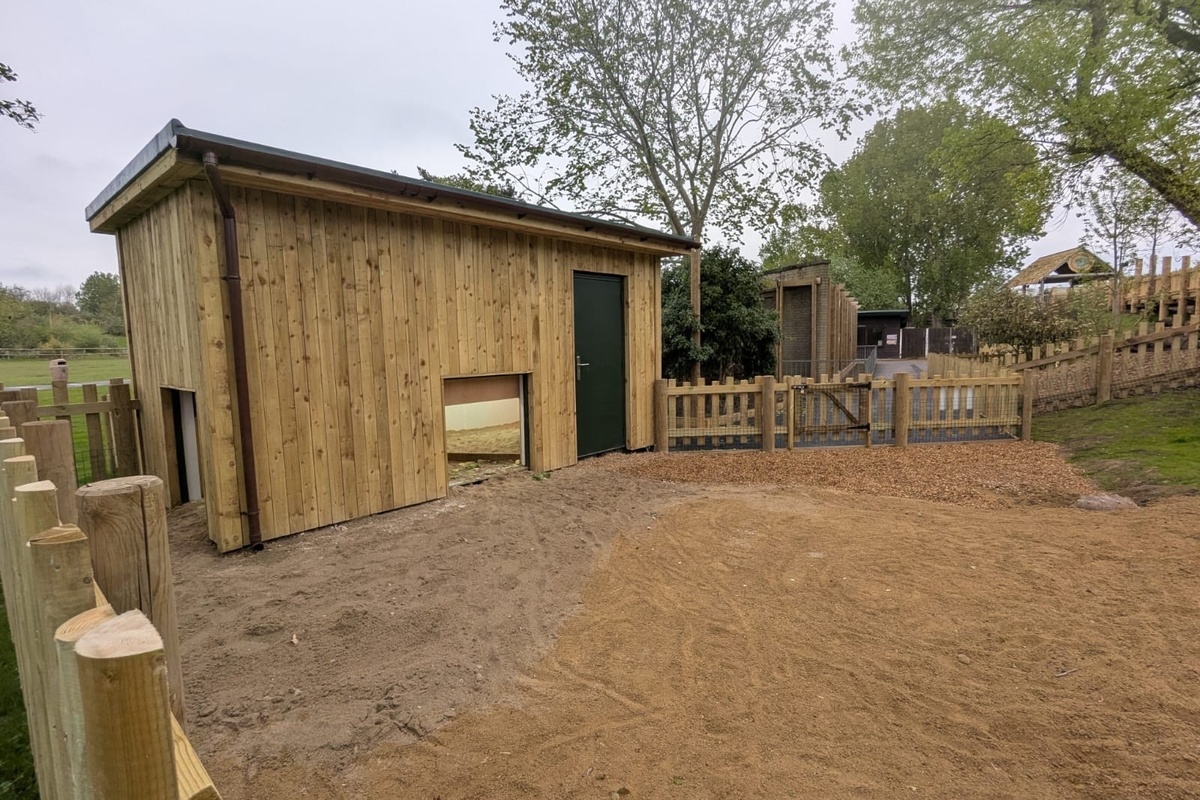 Wooden shed in a fenced area at Blackpool Zoo with trees and grass in the background.