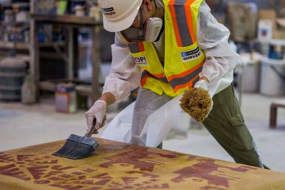 Worker paints stenciled wood, wearing safety gear and a neon safety vest in a workshop.