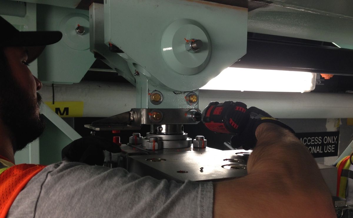 Worker uses a power drill on metal equipment under bright lighting.