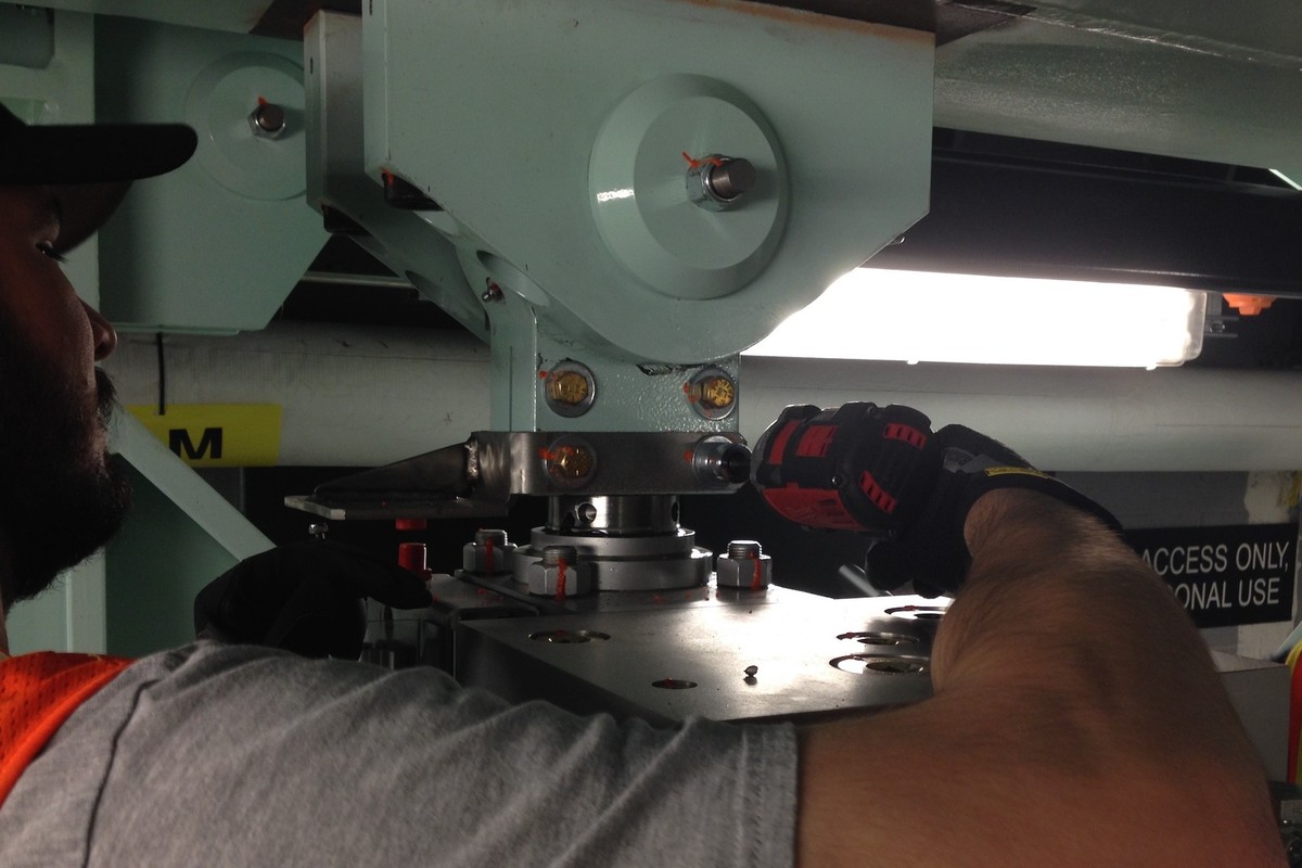 Worker uses a power drill on metal equipment under bright lighting.