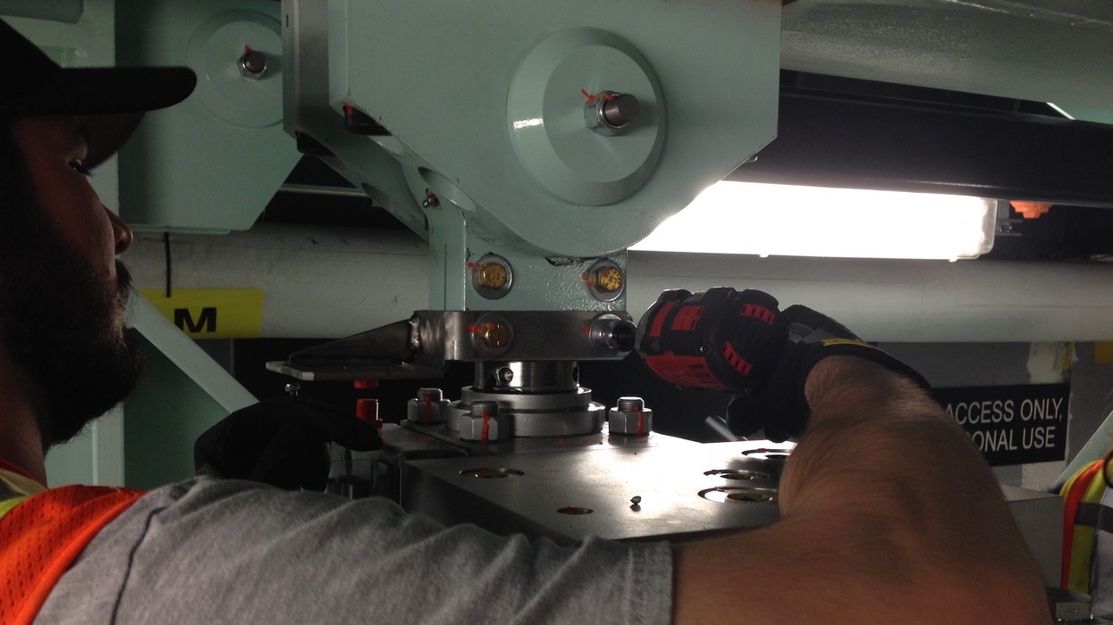 Worker uses a power drill on metal equipment under bright lighting.