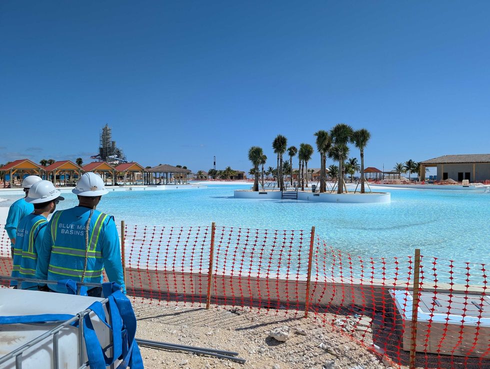 Workers in hard hats by a large blue pool with palm trees and buildings under construction.