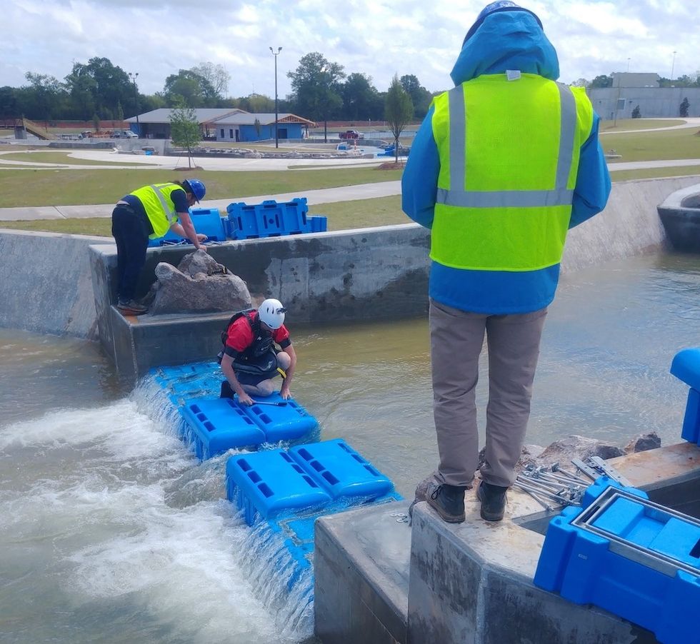 Workers in safety gear assemble blue blocks in a water channel at a construction site.