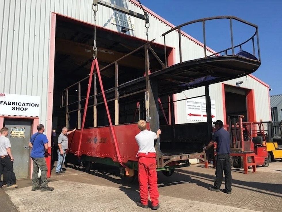 Workers lift a red tram body into a fabrication shop using a crane.