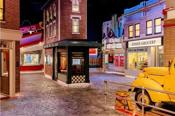 World of Coca-Cola: Coke Stories. Vintage street scene with a Coke sign, radio store, grocery, and yellow truck.